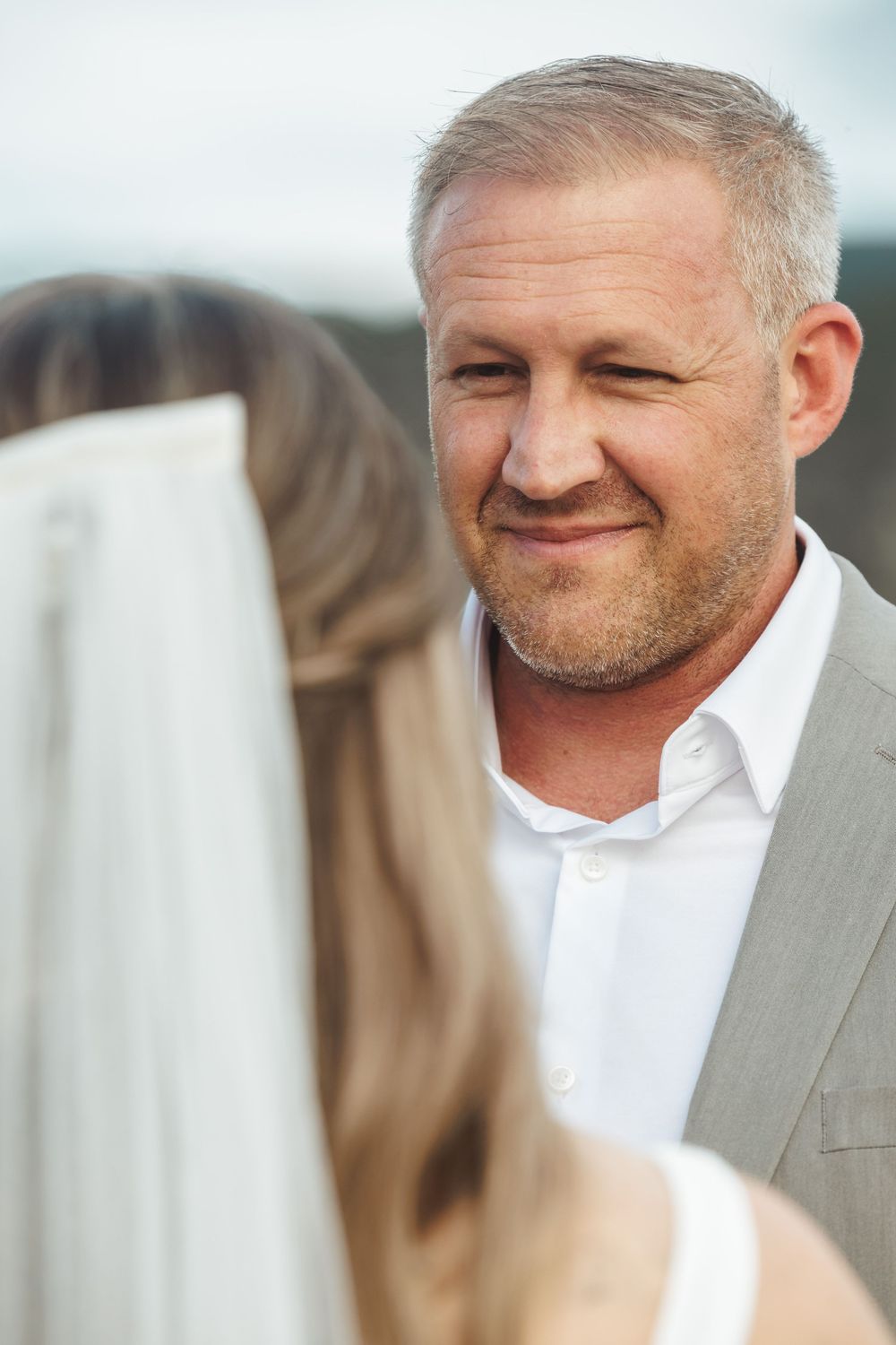 Groom smiling warmly at bride during outdoor wedding ceremony with mountain backdrop.