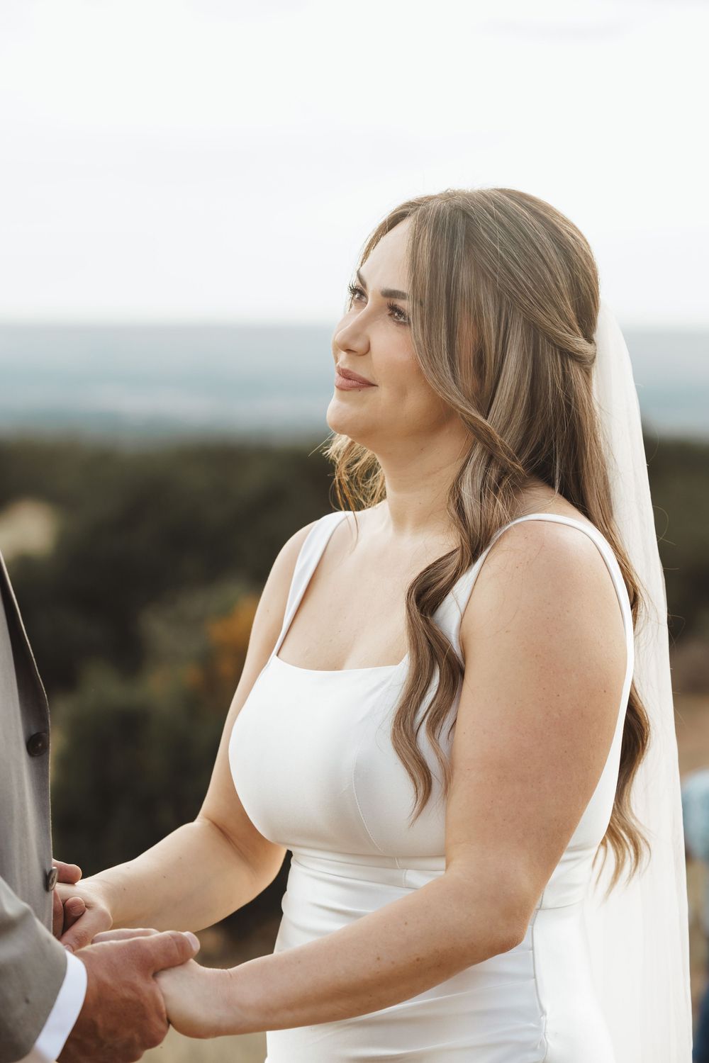 Bride in white sleeveless gown and veil holds hands during outdoor wedding ceremony with scenic landscape backdrop.