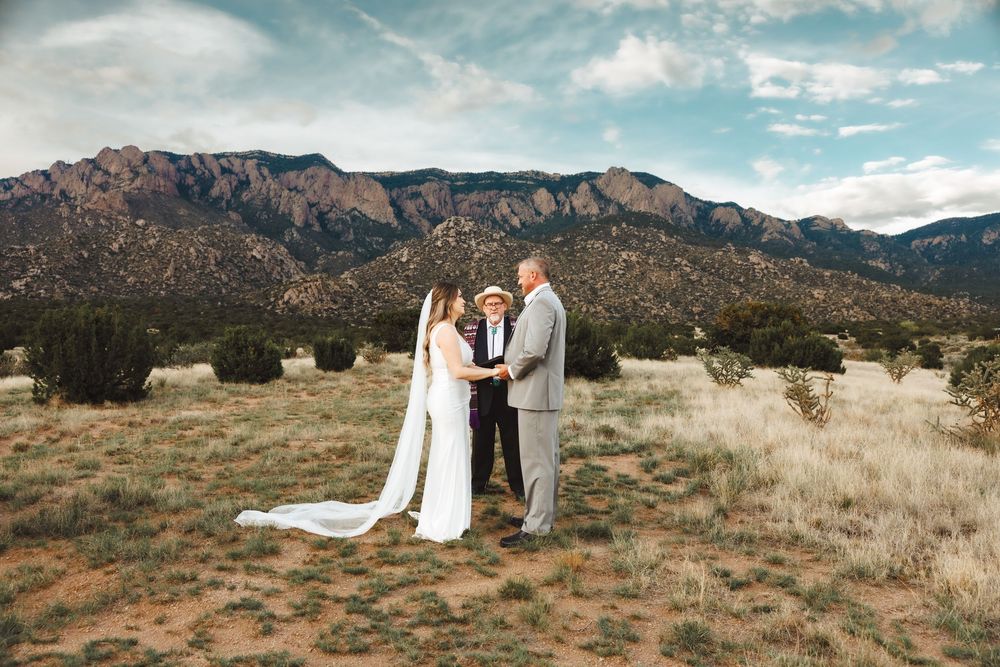 Outdoor wedding ceremony in New Mexico desert with mountains, bride in white gown, groom, and officiant holding hands.