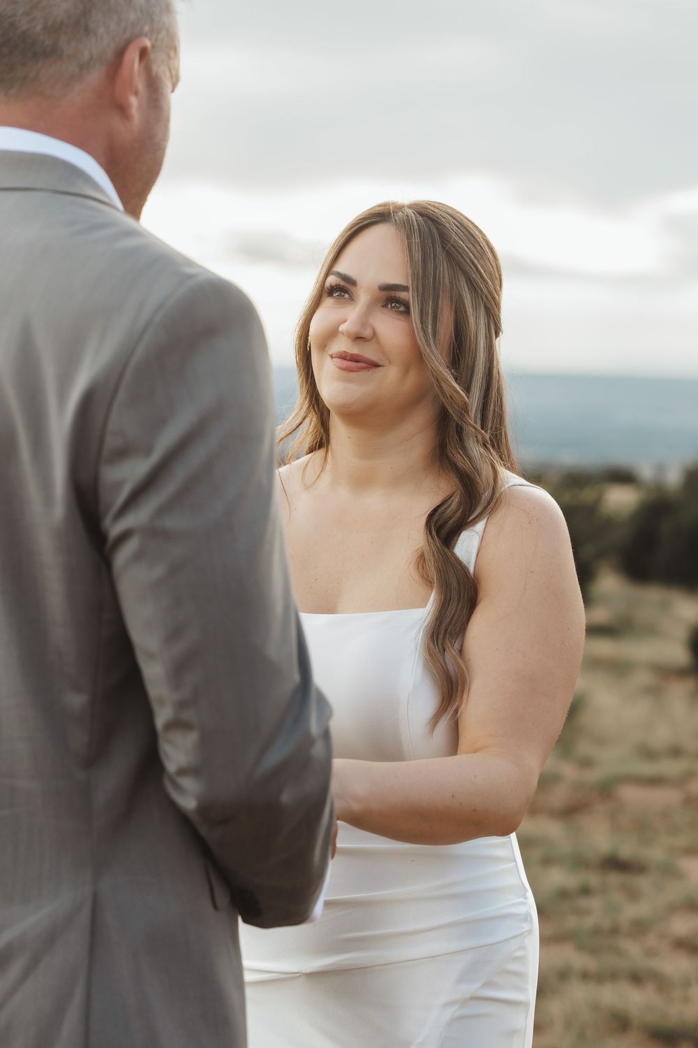 Bride in white strapless gown holds hands with groom in gray suit during outdoor wedding ceremony.