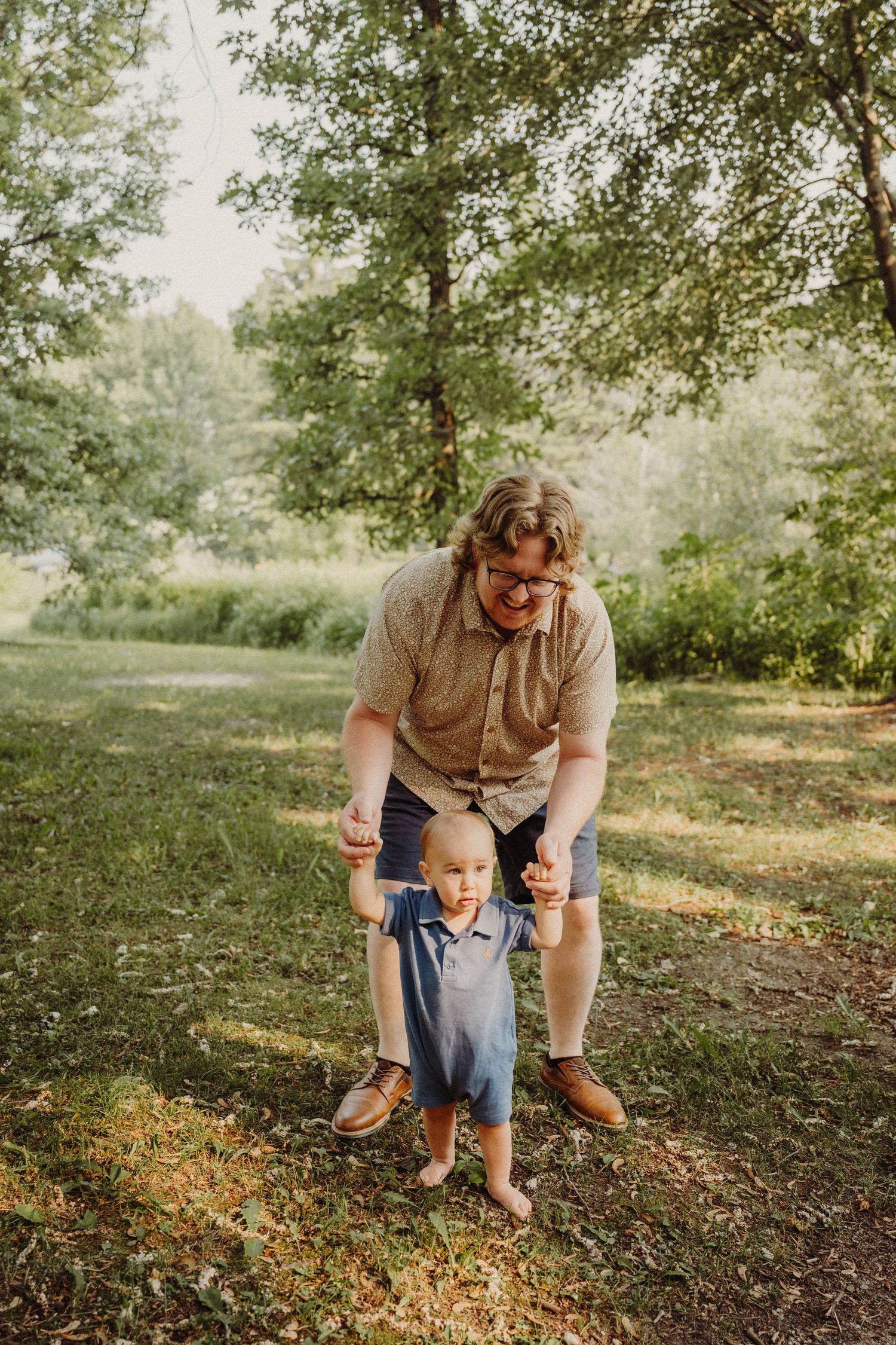 Father helps barefoot toddler take first steps outdoors in lush green park on sunny summer day.