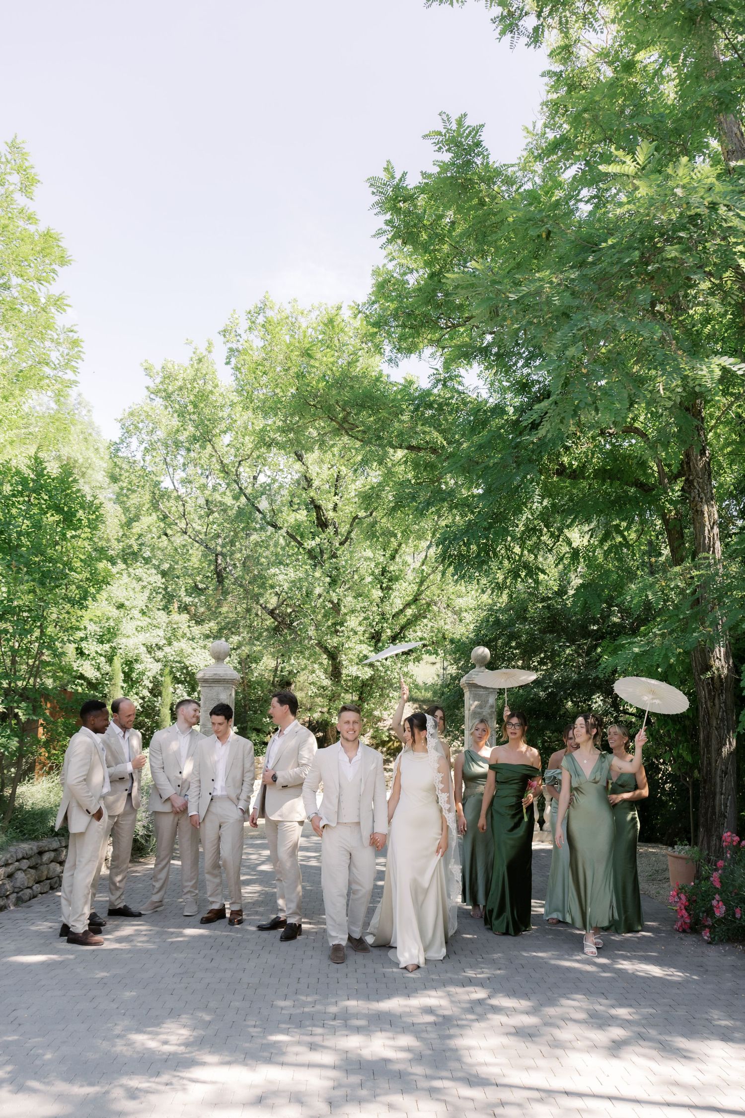 Wedding party walks outdoors in lush greenery, groomsmen in tan suits, bridesmaids in green gowns holding parasols.