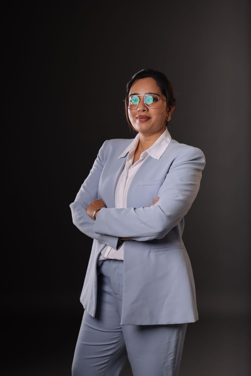 Professional woman in light blue suit with arms crossed, wearing green glasses, posing confidently against dark background.