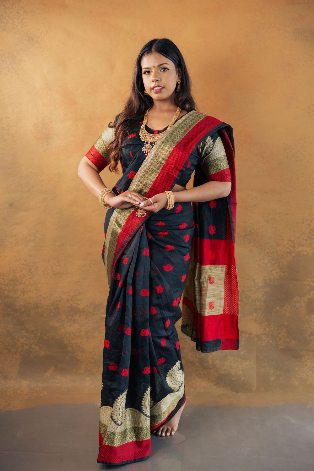 Woman in black and red Banarasi silk saree with gold jewelry posing gracefully against a golden background.