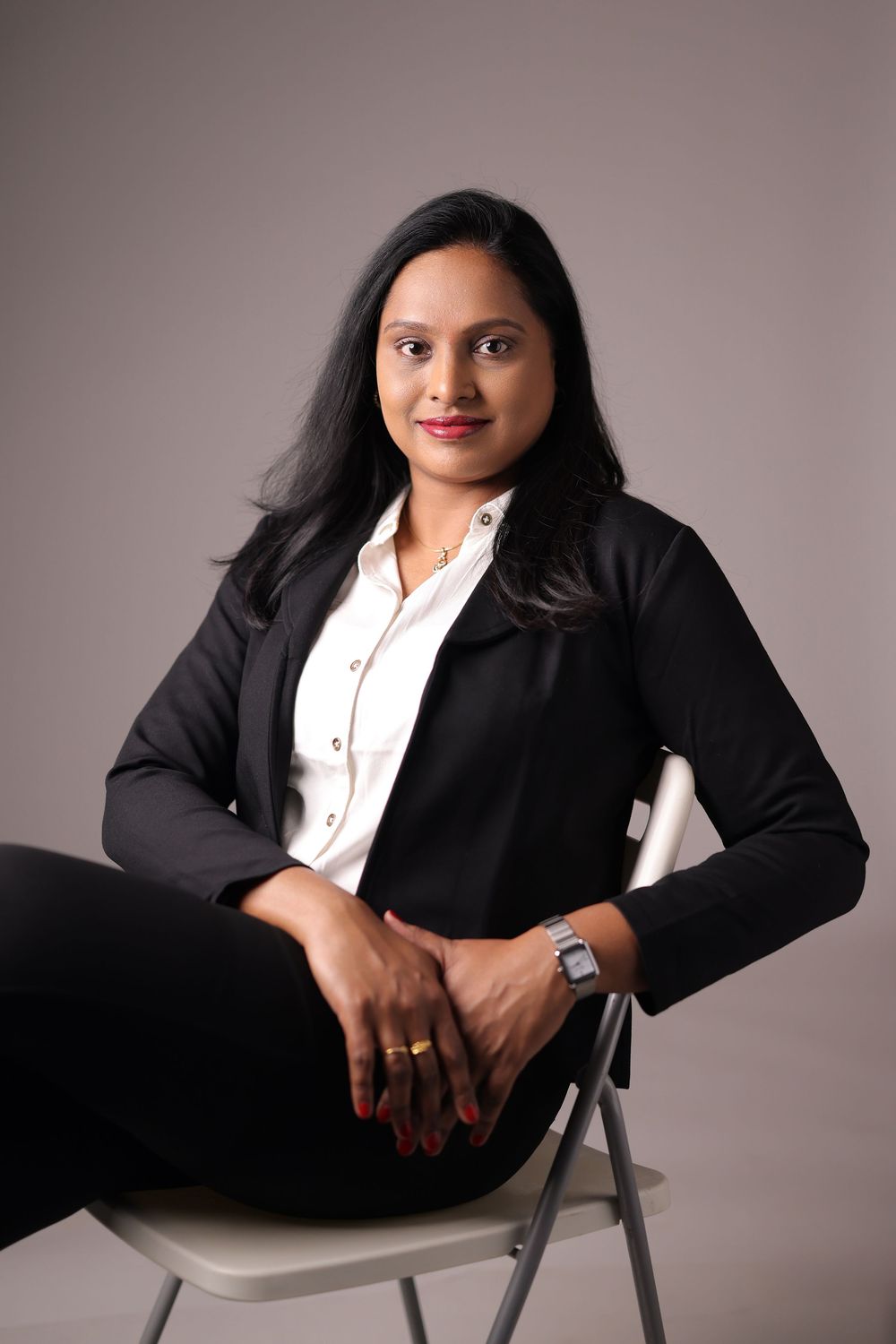 Professional woman in black blazer and white shirt seated on chair, smiling confidently in studio portrait.