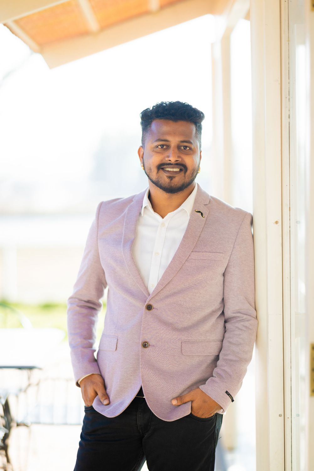 Smiling man in pink blazer and white shirt standing confidently with hands in pockets outdoors.