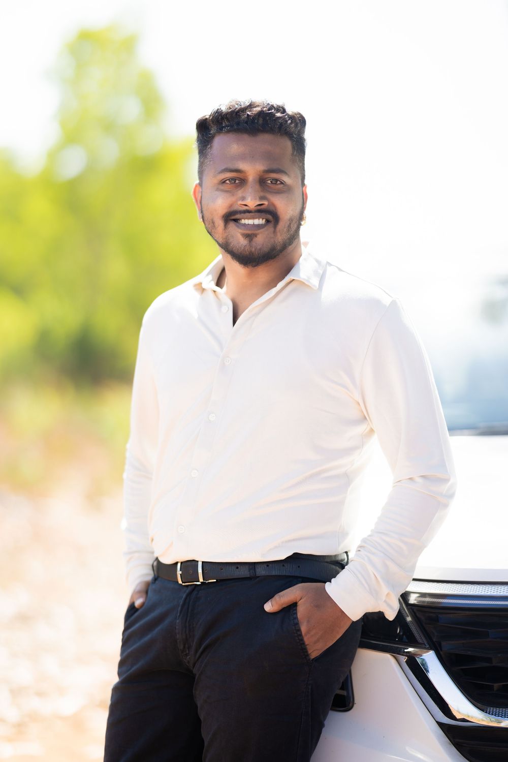 Smiling Indian man in white shirt and black pants leaning against a white car outdoors.