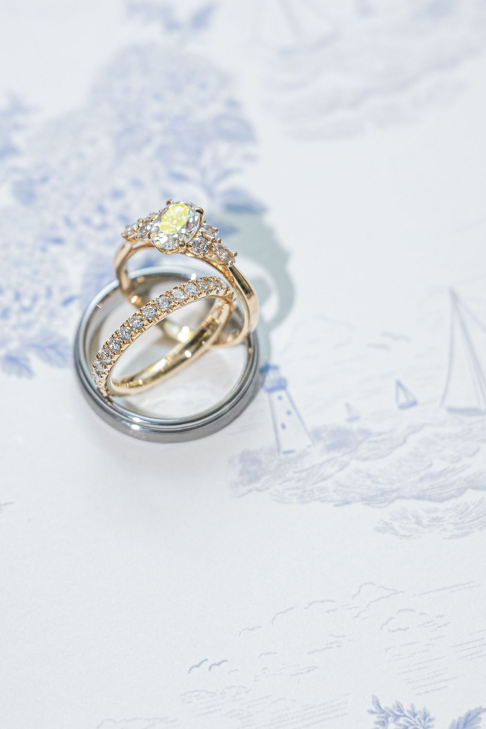 Three elegant wedding rings stacked together, featuring a diamond halo engagement ring in rose gold on a blue toile background.