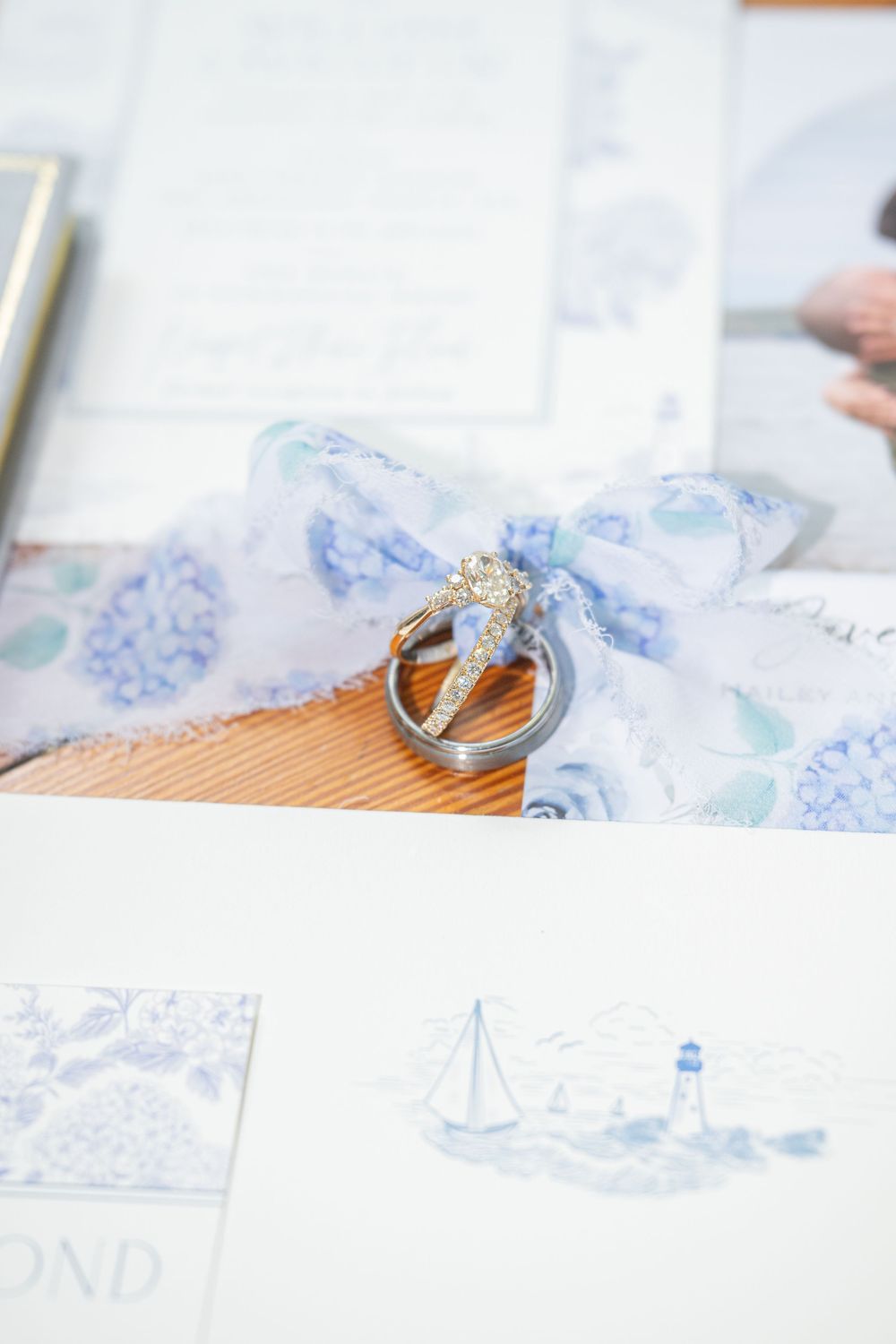 Wedding rings resting on blue hydrangea ribbon beside nautical-themed stationery with sailboat and lighthouse illustrations.