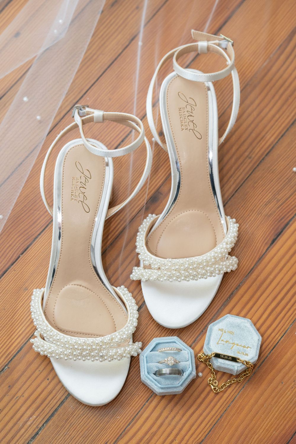 White pearl-embellished bridal heels with ankle straps displayed on wood floor beside a blue velvet ring box with diamond rings.