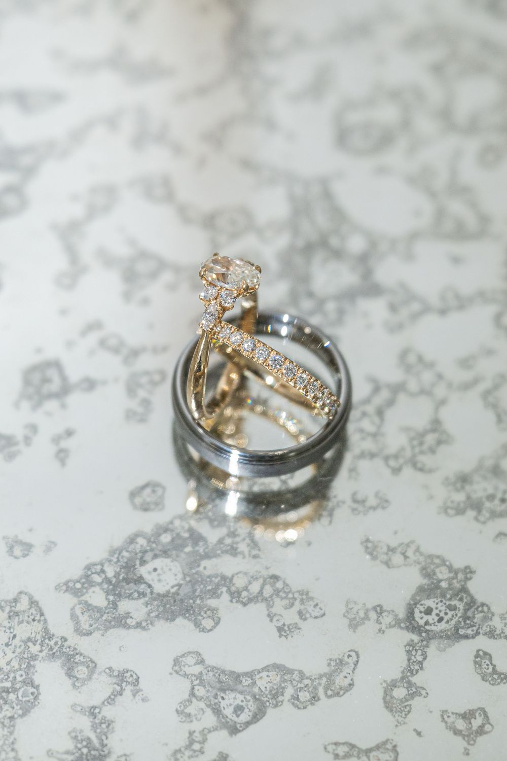 Gold diamond engagement ring and silver wedding bands stacked together on a marble surface.