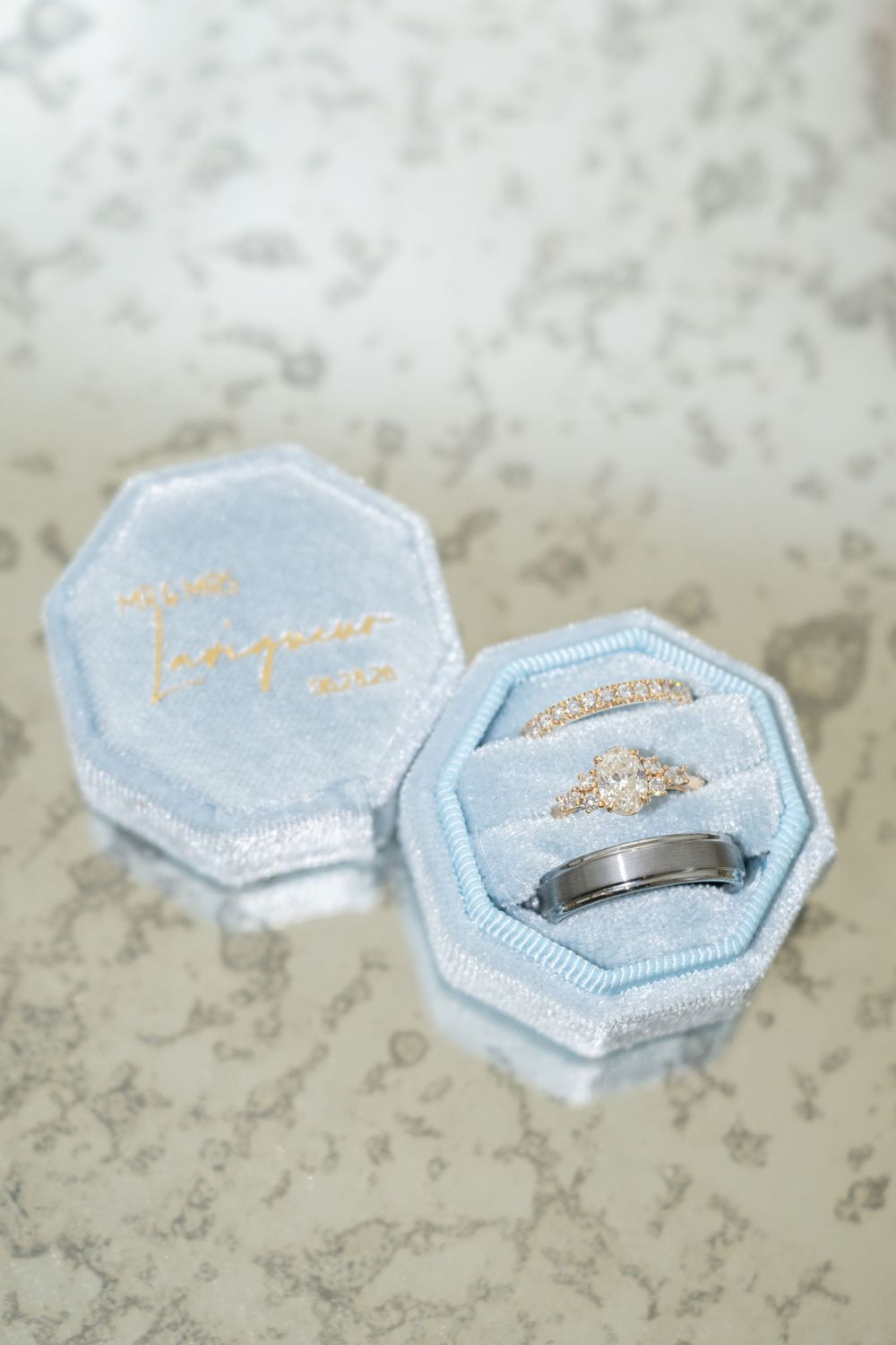Light blue velvet octagonal ring box displaying diamond engagement ring, wedding band, and mens ring on soft blue cushion.