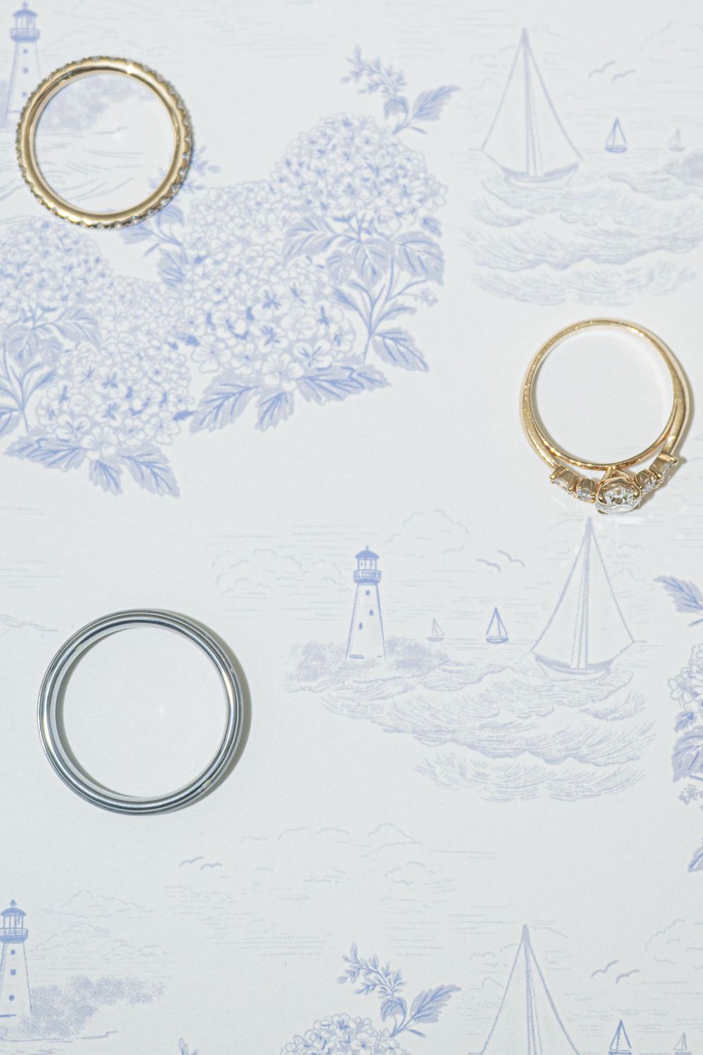 Gold engagement ring, gold band, and silver band displayed on blue nautical toile wallpaper with lighthouse and sailboat motifs.