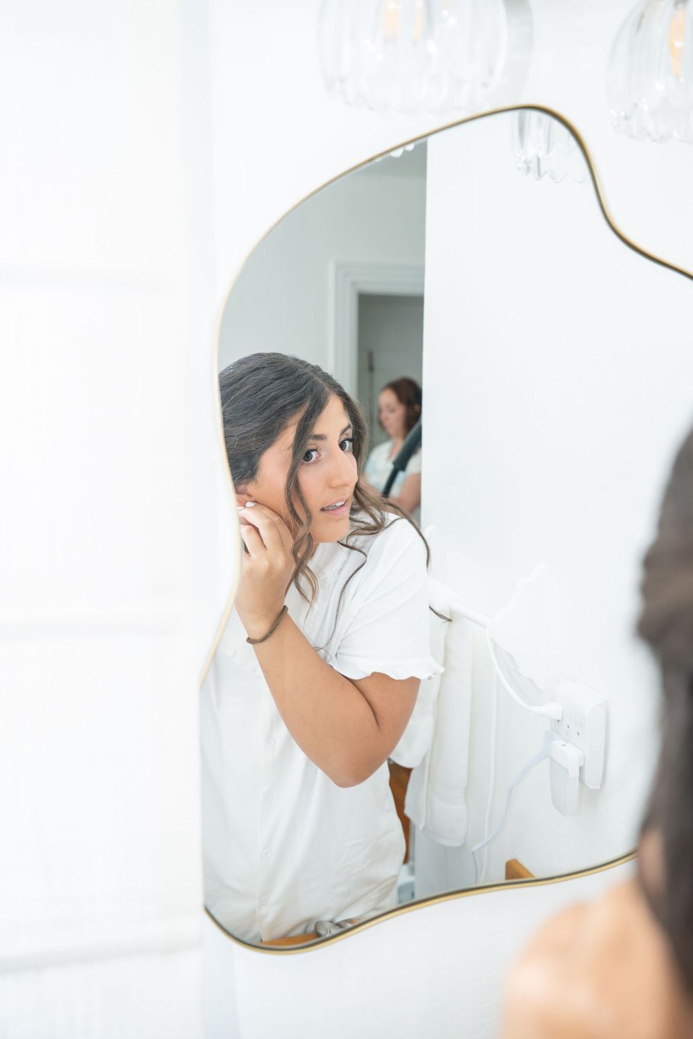 Bride in white robe adjusting earring while looking at her reflection in an ornate gold-framed mirror.