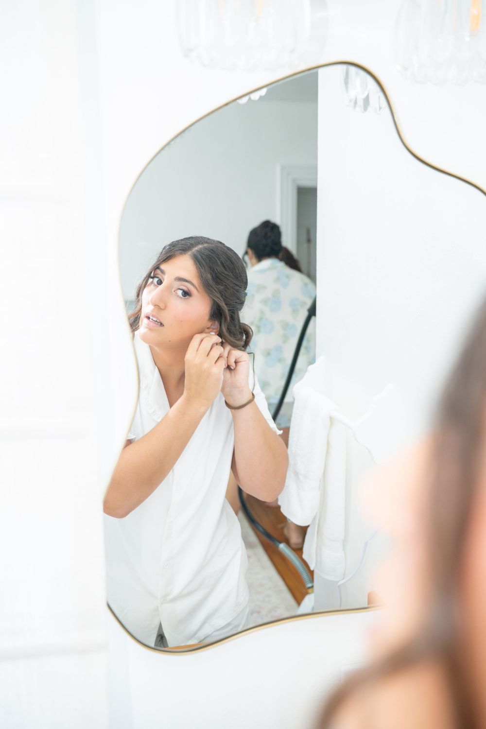 Bride in white robe putting on earrings while looking in an ornate gold-framed mirror during wedding preparations.
