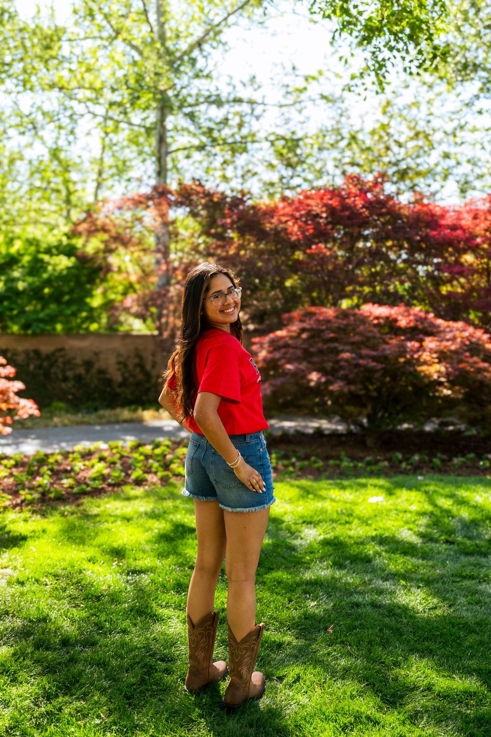 Young woman in red top, denim shorts, and cowboy boots smiling in a lush green garden with colorful foliage.