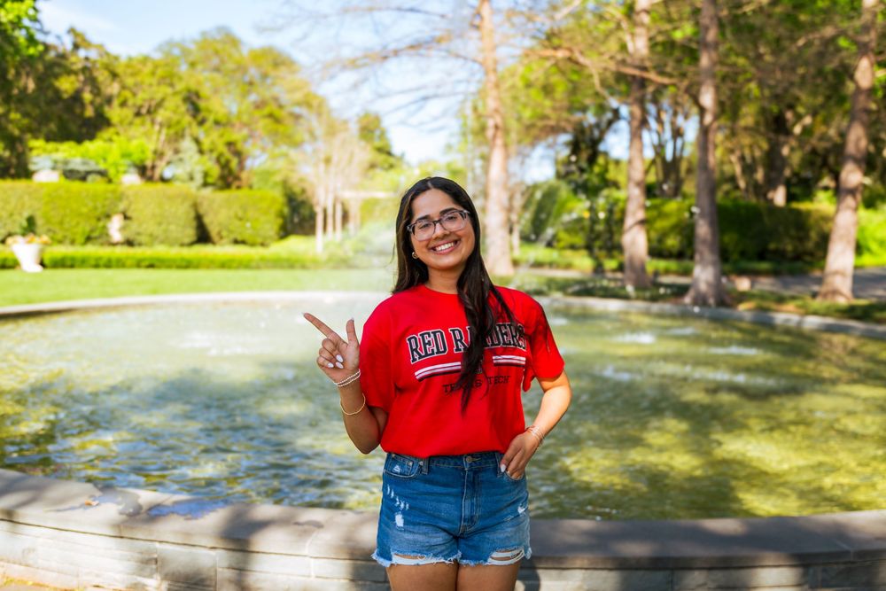Young woman in red college shirt and denim shorts smiling by a fountain in a sunny park.