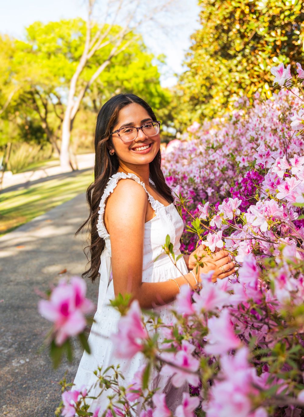 Young woman in white dress smiling among pink azalea blooms in a sunny park setting.