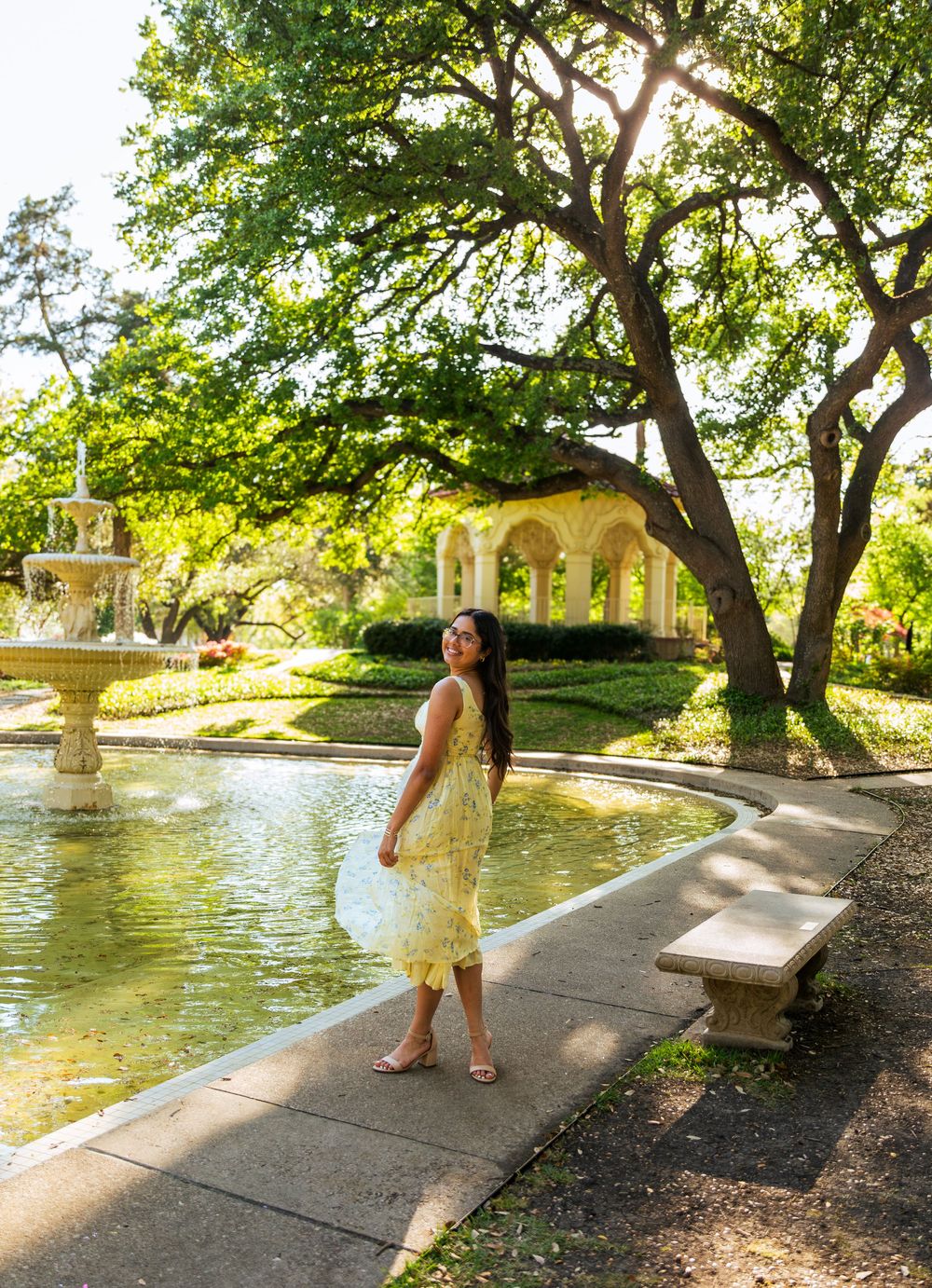 Woman in yellow floral dress posing by a garden fountain with lush trees and a classical pergola in the background.