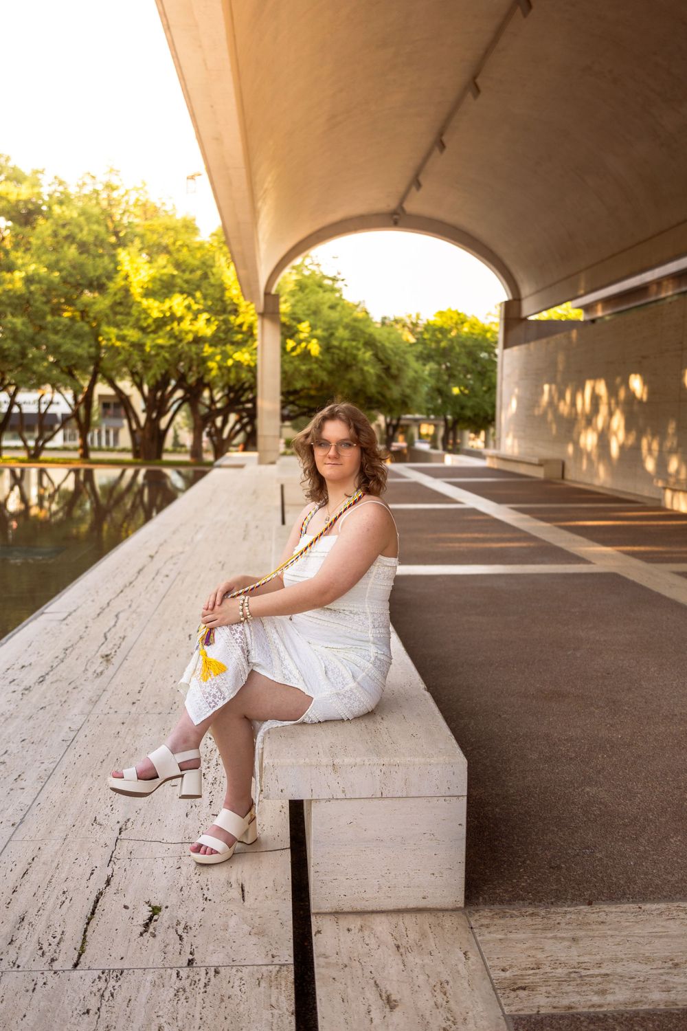 Woman in white dress sitting on concrete bench under arched structure with green trees in background.