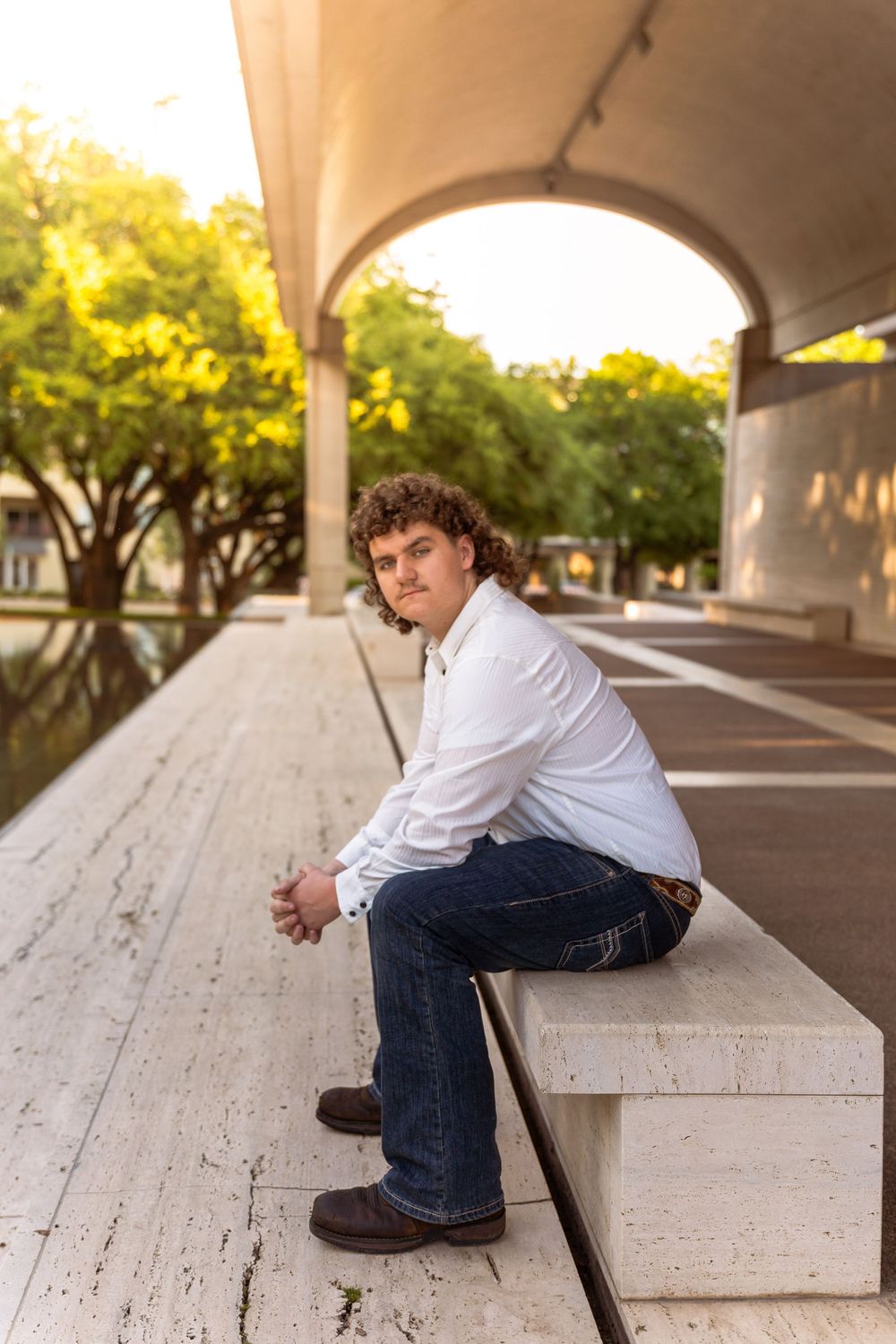 Young man with curly hair in white shirt and jeans sits on stone steps under arched colonnade at golden hour.