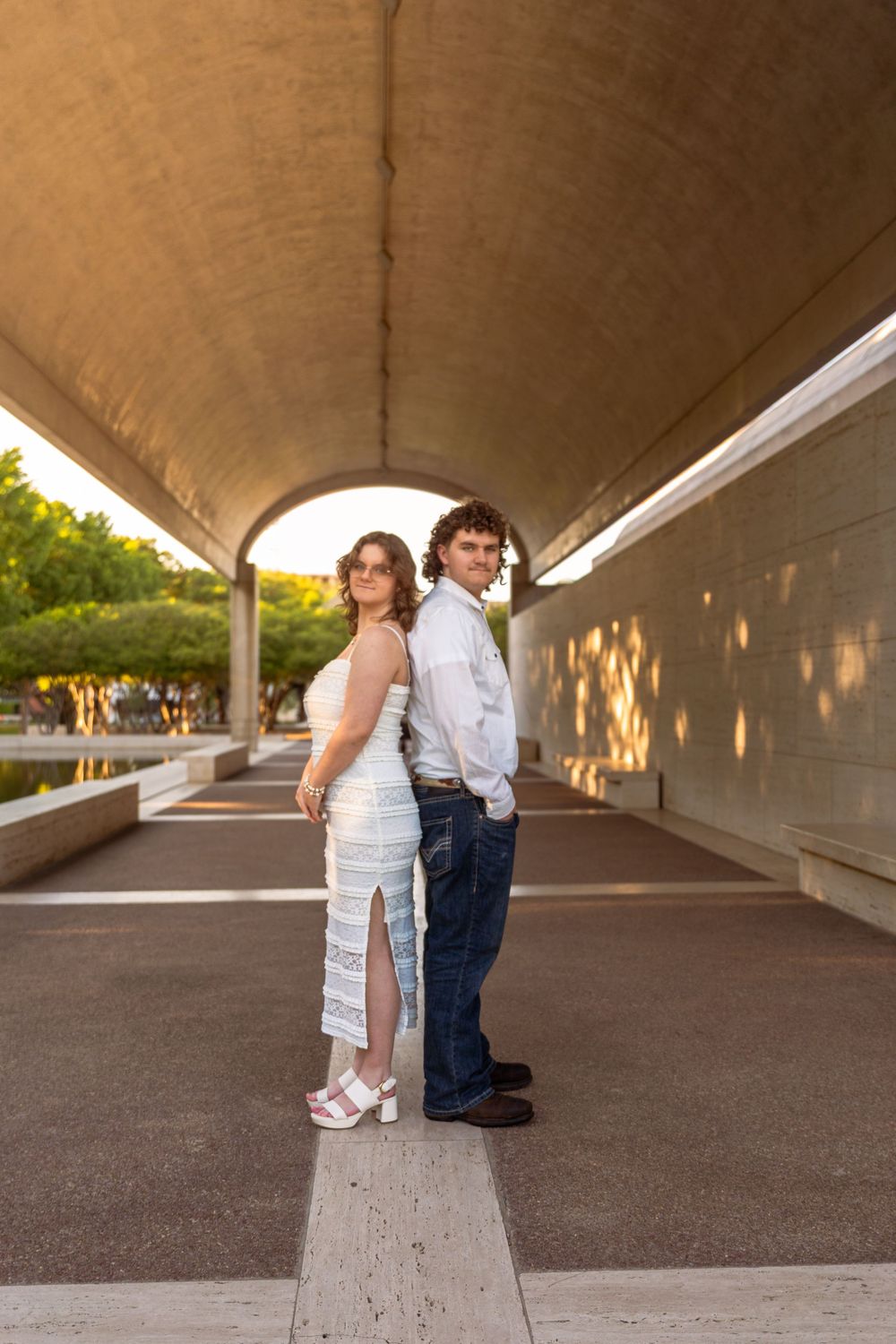 Couple posing back-to-back under an arched tunnel walkway, woman in white dress and man in jeans and white shirt.