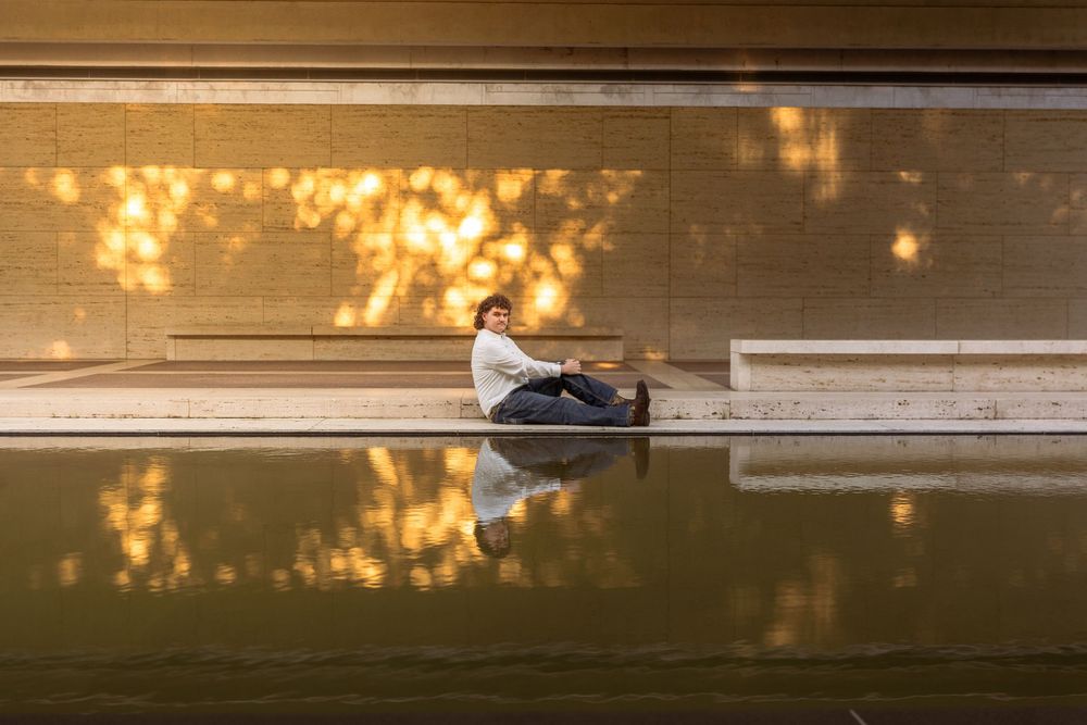 Person sitting beside a reflective pool with golden light and tree shadows on a warm stone wall.