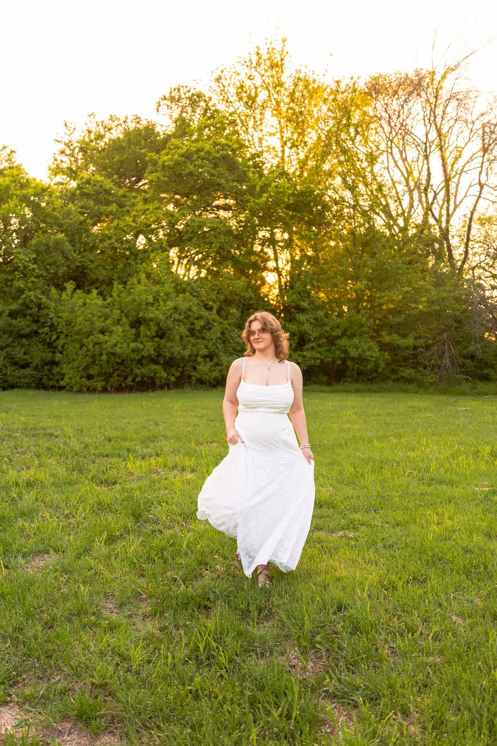 Woman in white dress walking through green field at golden hour sunset with trees in background.