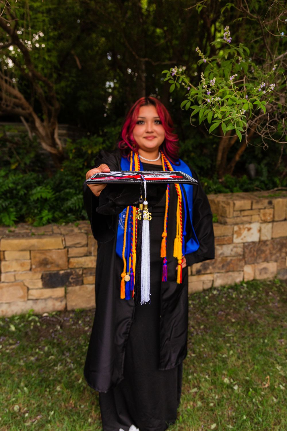 Young woman with red hair in black graduation gown holding cap, wearing colorful honor cords outdoors.