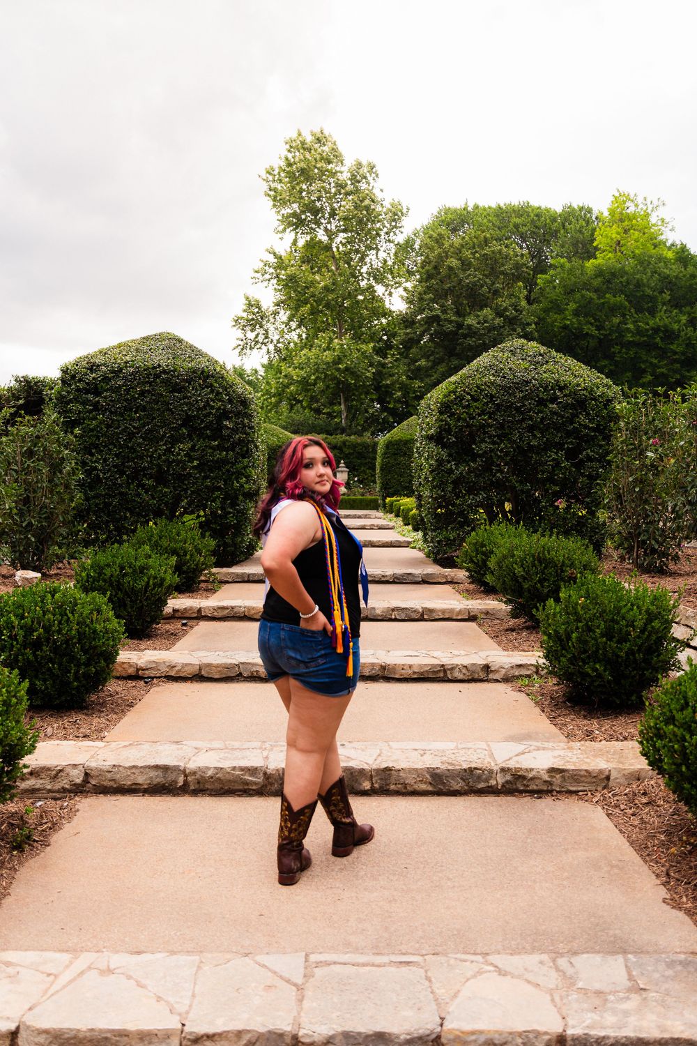 Woman with red hair in denim shorts and boots poses on stone path in manicured garden with trimmed hedges.