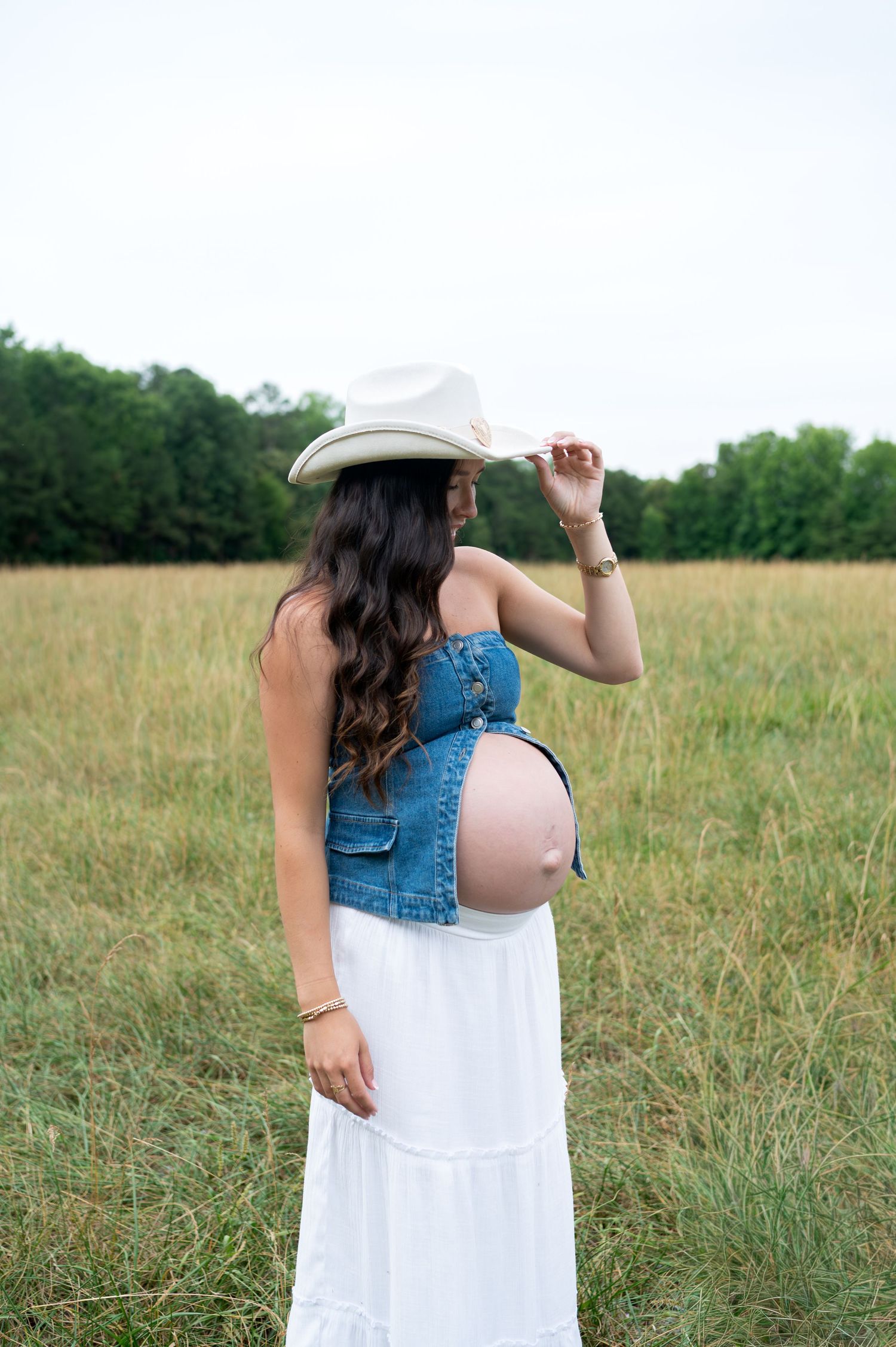 Pregnant woman in denim overalls and cowboy hat standing in an open field, maternity photographer NC