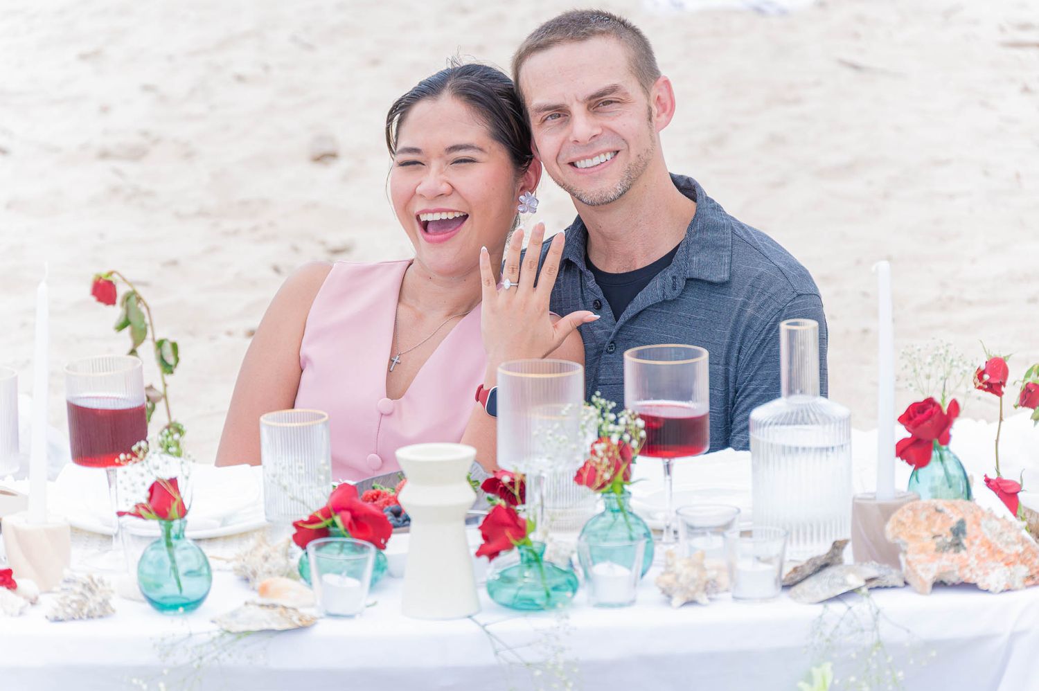 Luxury proposal decorated beach table showing engagement ring, smiling joyfully with floral arrangements and candles around them.