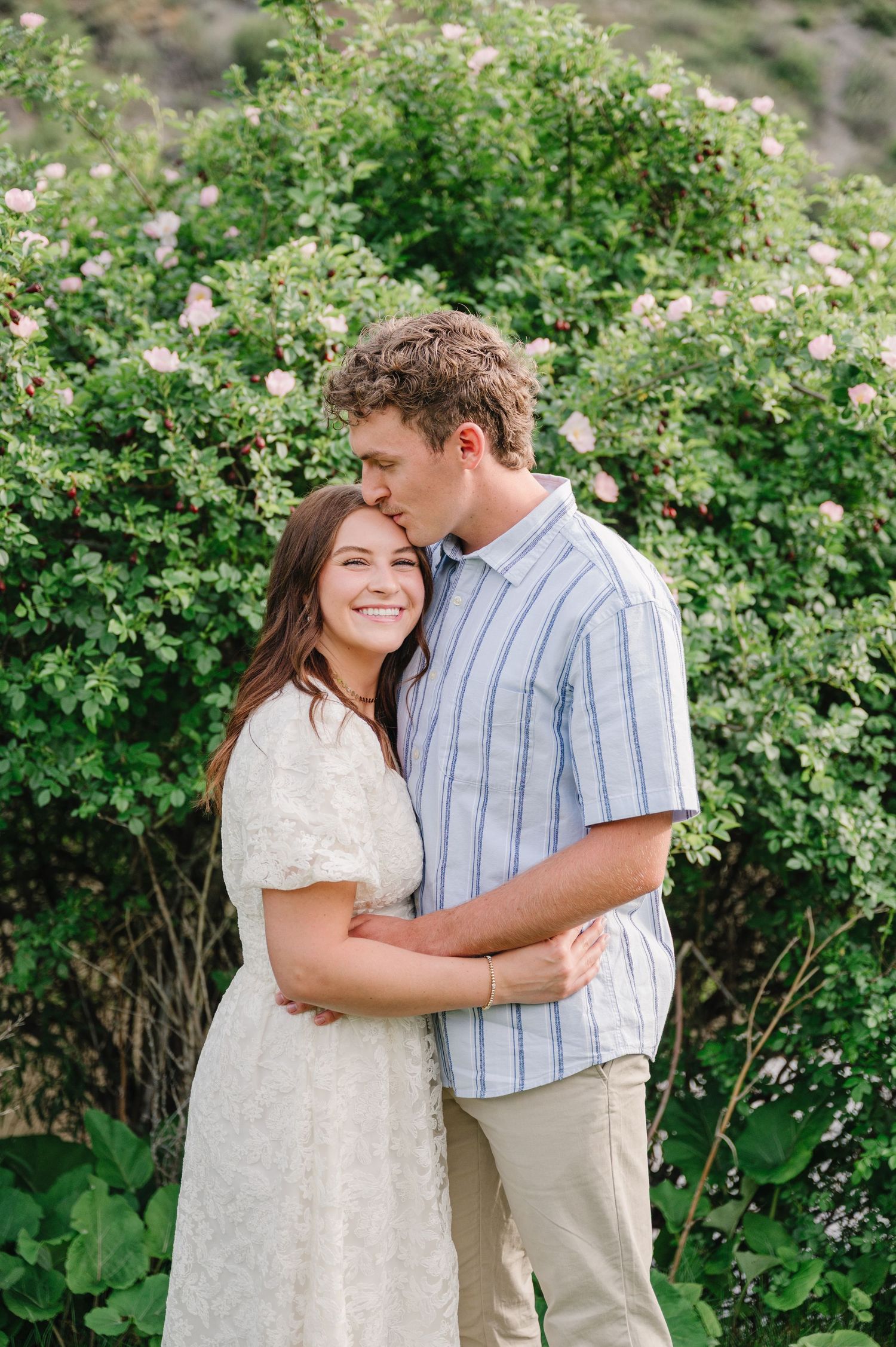 A couple embraces by blooming bushes, with her in a white dress and him in a striped shirt.