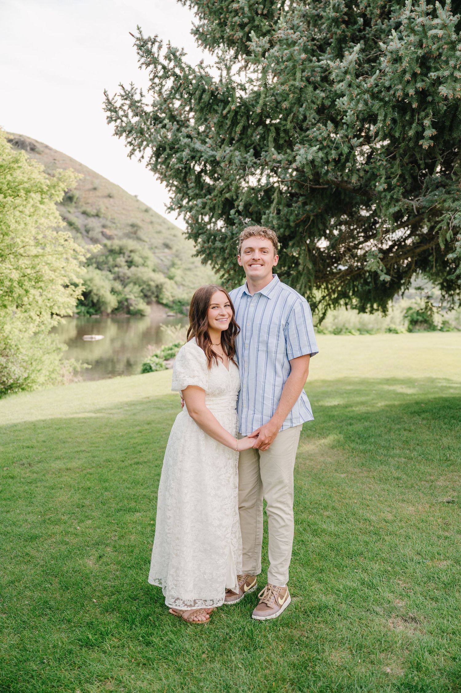 A couple stands together on green grass near a lake and trees in a natural outdoor setting.