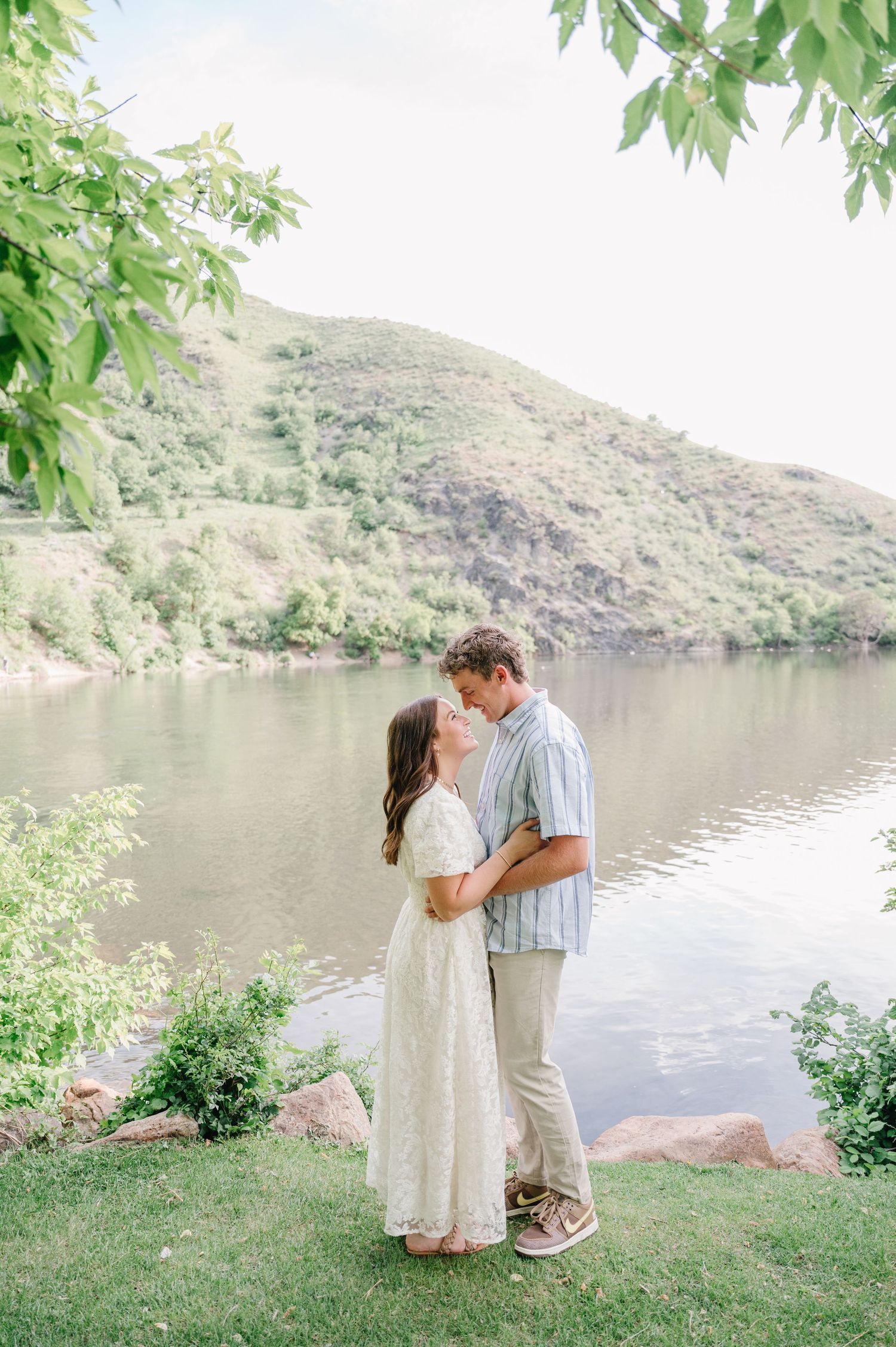 Romantic couple embraces by a serene mountain lake surrounded by lush greenery on a sunny day.