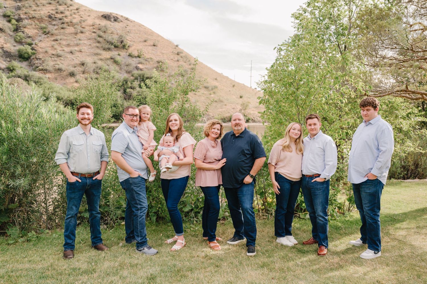 Large family group portrait outdoors with green hills, wearing coordinated pink and blue outfits on a sunny day.