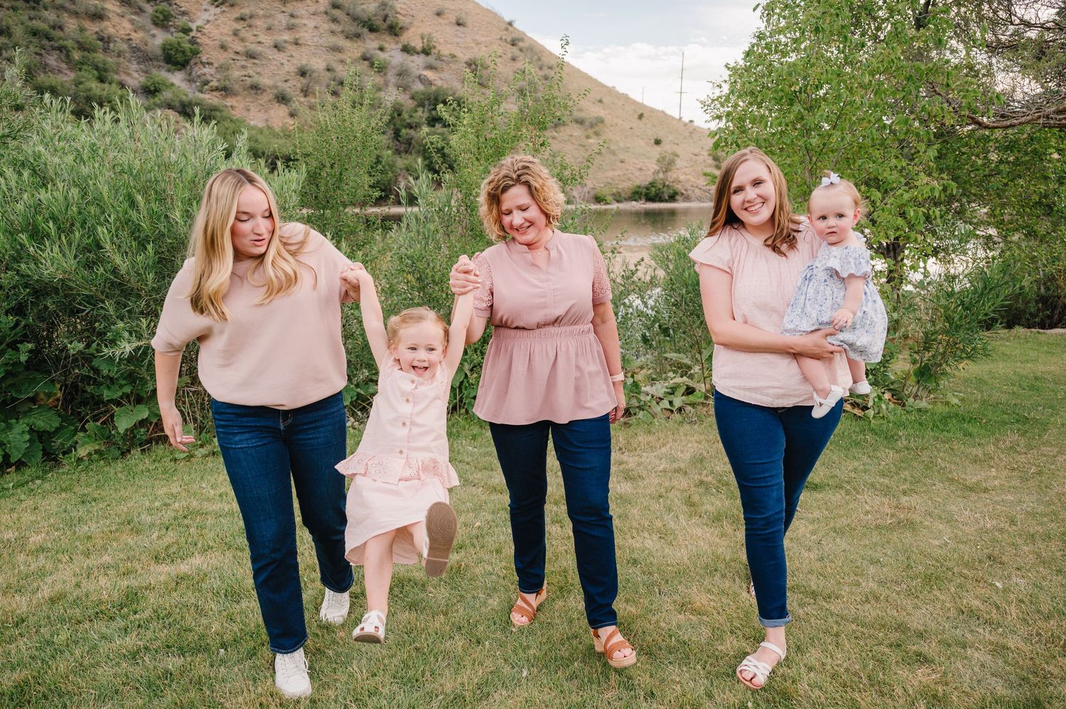 Three women in pink tops and jeans walk outdoors with two toddlers near a scenic lake and green hillside.