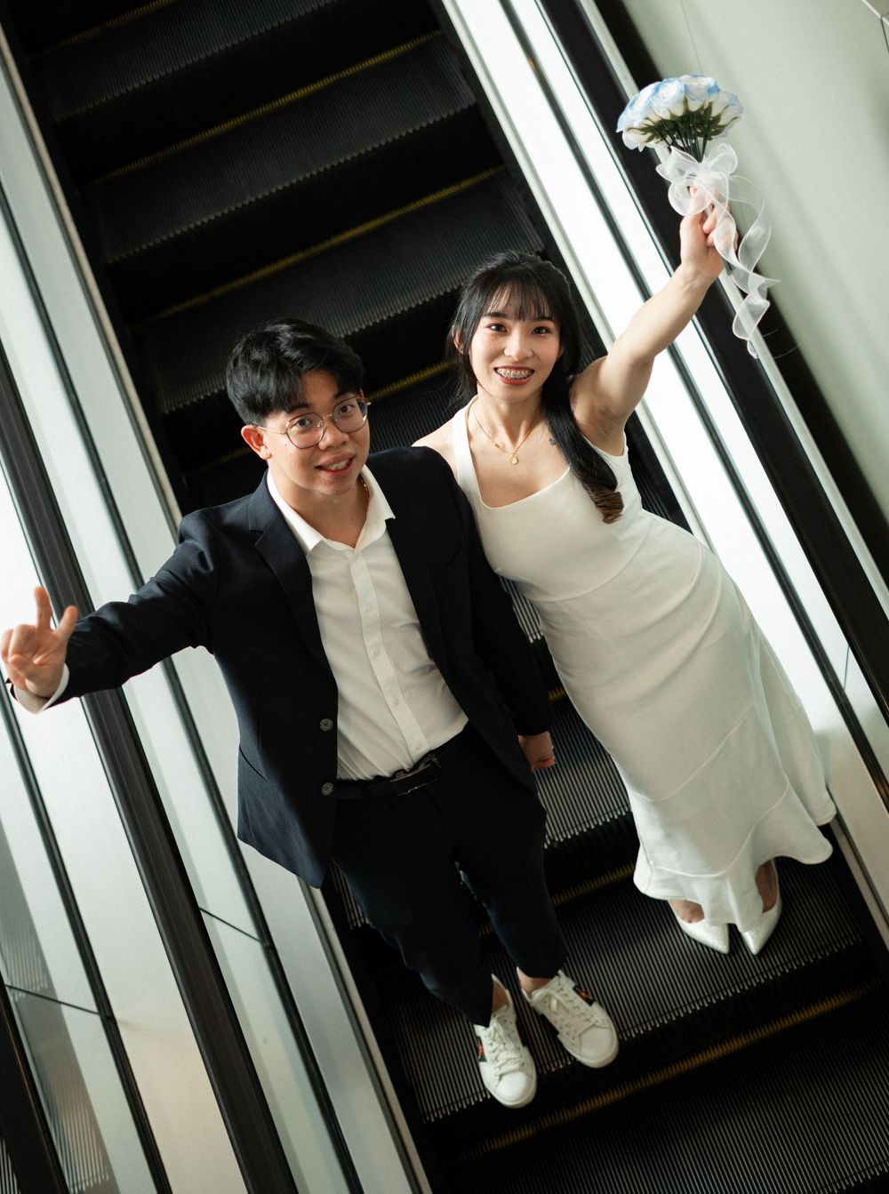 Happy couple in wedding attire celebrating on an escalator, bride holding bouquet in white dress, groom in black suit.