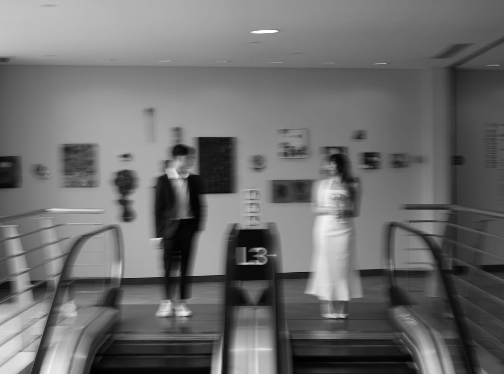 Black and white photo of a man and woman standing on an escalator in a modern art gallery.