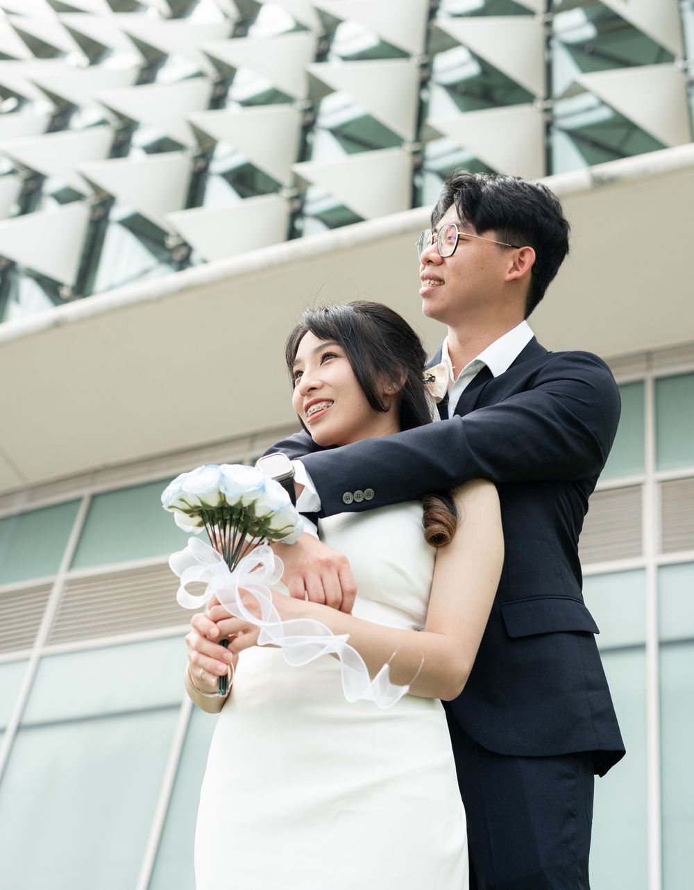 A couple in wedding attire poses outdoors, the groom in a suit embracing the bride holding a white bouquet.