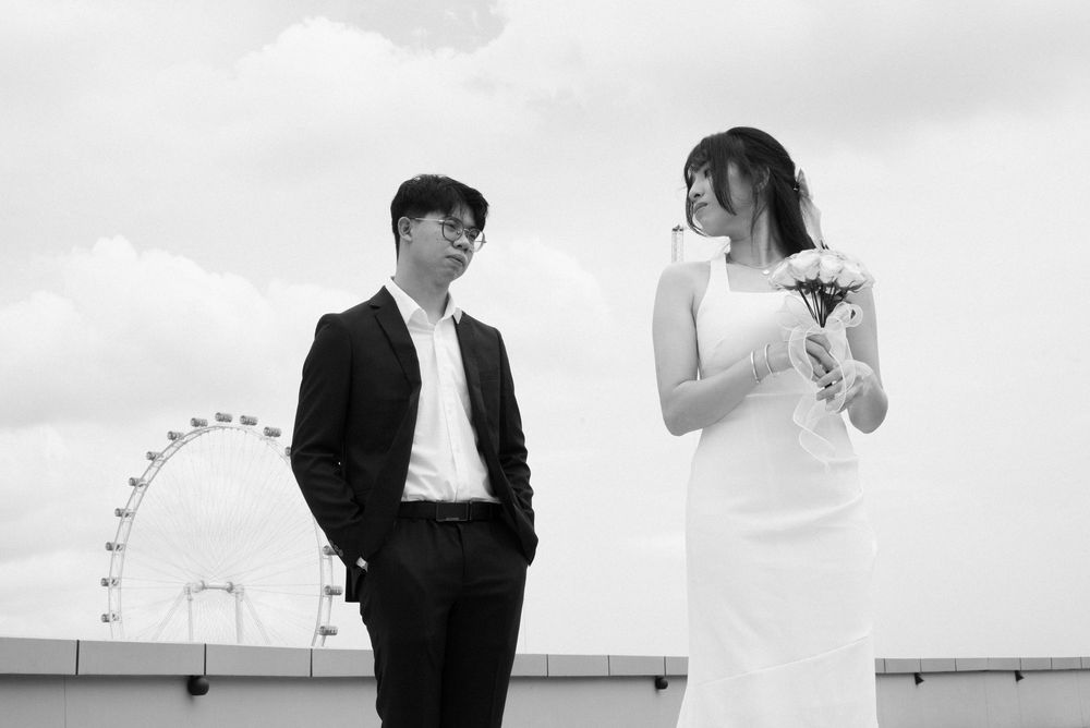 Black and white photo of a couple in formal wear near a Ferris wheel, man in suit, woman in white dress holding flowers.