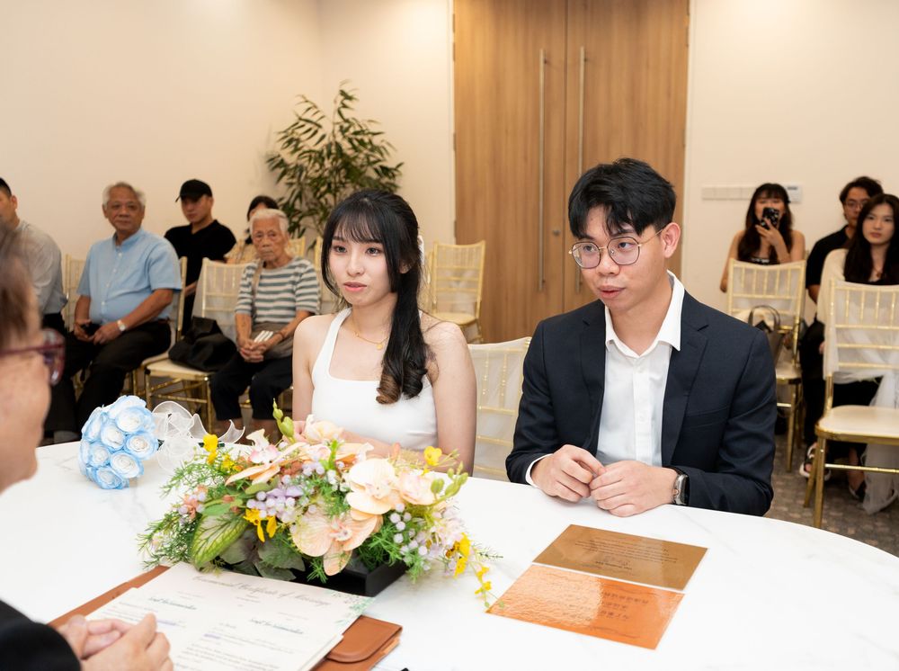 Young Asian couple sits at a table during a formal ceremony, with guests seated in the background.