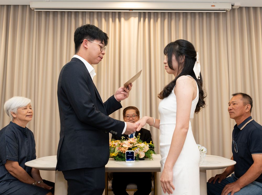 A groom reads vows to his bride in white dress during an intimate indoor wedding ceremony with guests seated nearby.