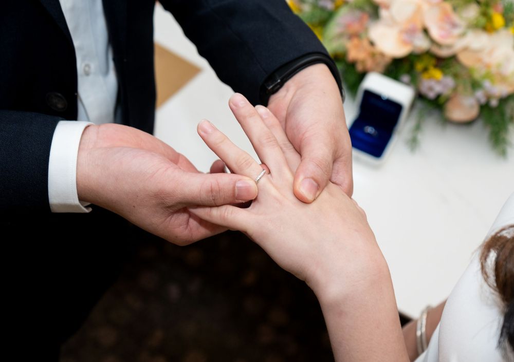 Groom placing a wedding ring on bride's finger during a romantic ceremony with flowers in the background.