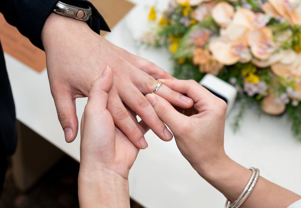 Close-up of wedding ring exchange ceremony with floral arrangement in background.