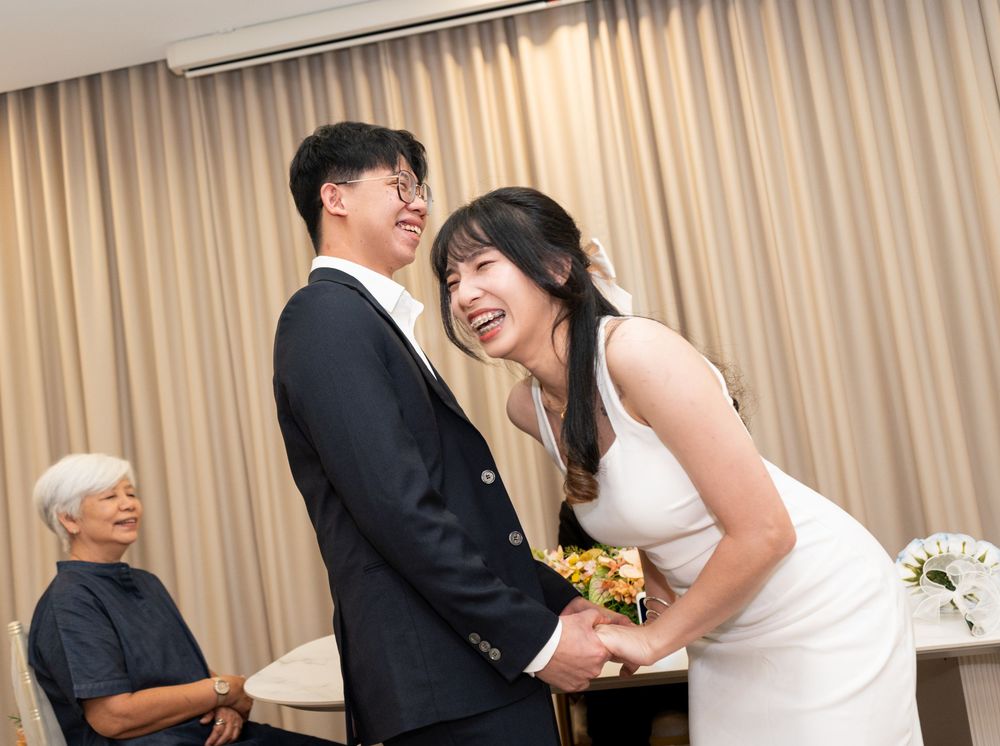 A joyful couple in wedding attire laughs together, the bride holding yellow flowers, with an elderly man seated in the background.
