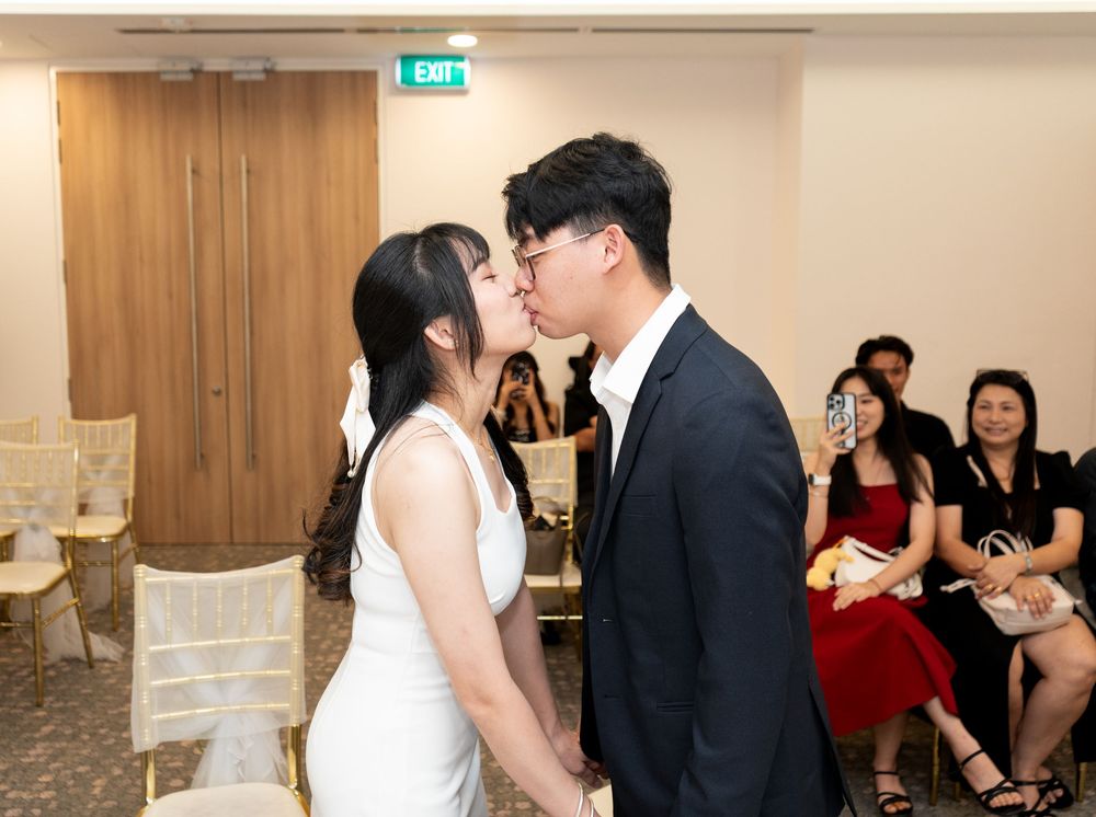 Couple sharing a first kiss at an indoor wedding ceremony with guests seated in the background.