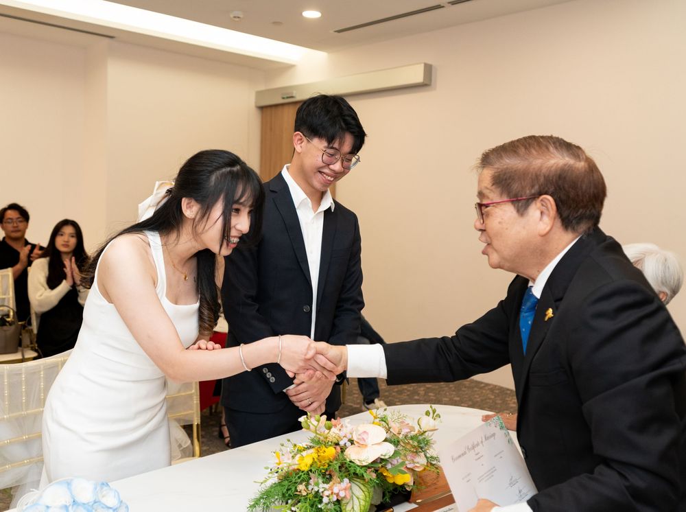 A bride in a white dress shakes hands with a suited official at a wedding reception ceremony.