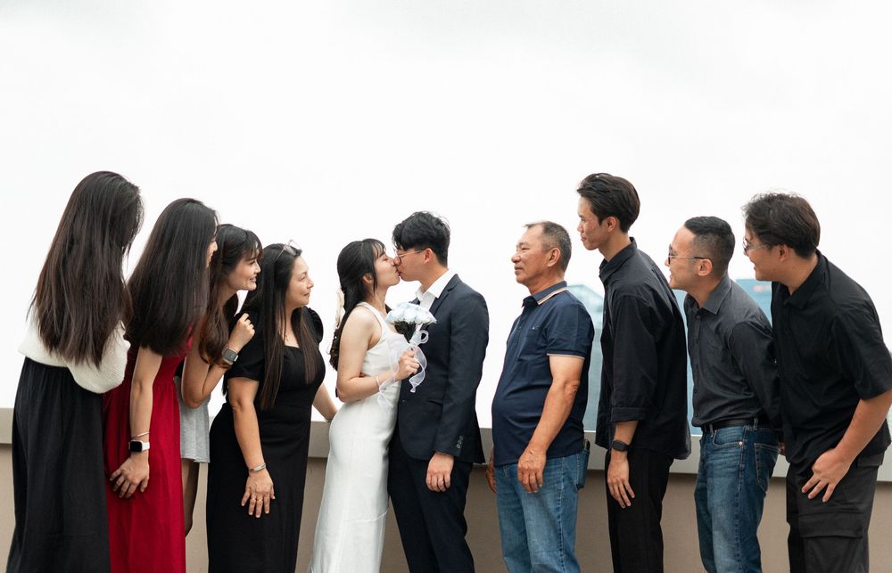 Wedding couple kissing surrounded by smiling guests on rooftop, group photo with bridesmaids and groomsmen.