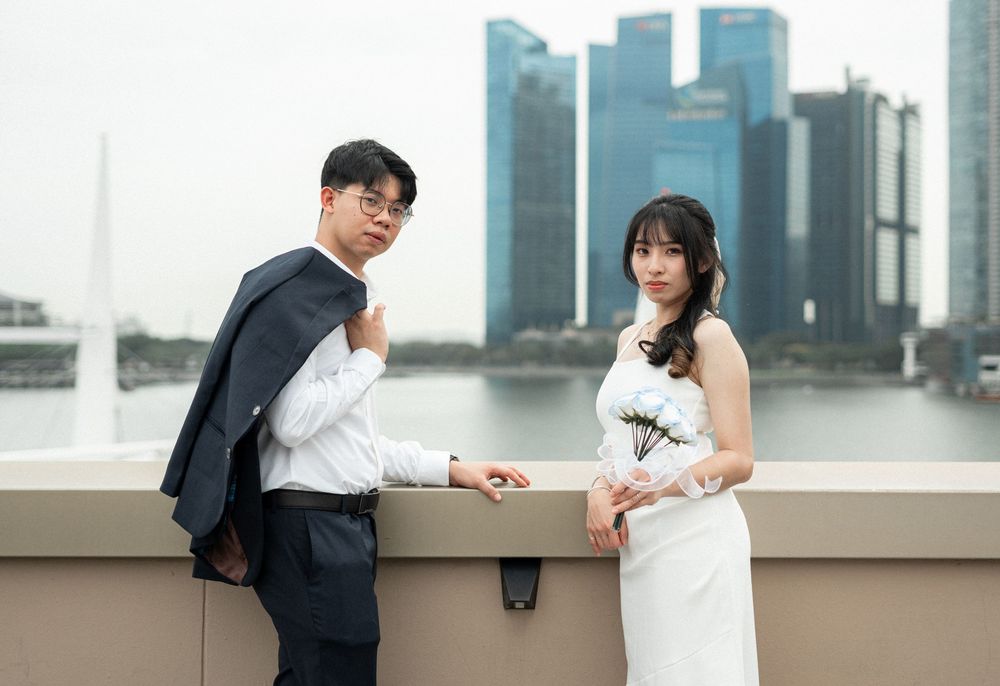 Couple in wedding attire posing on waterfront with Singapore city skyline in background.